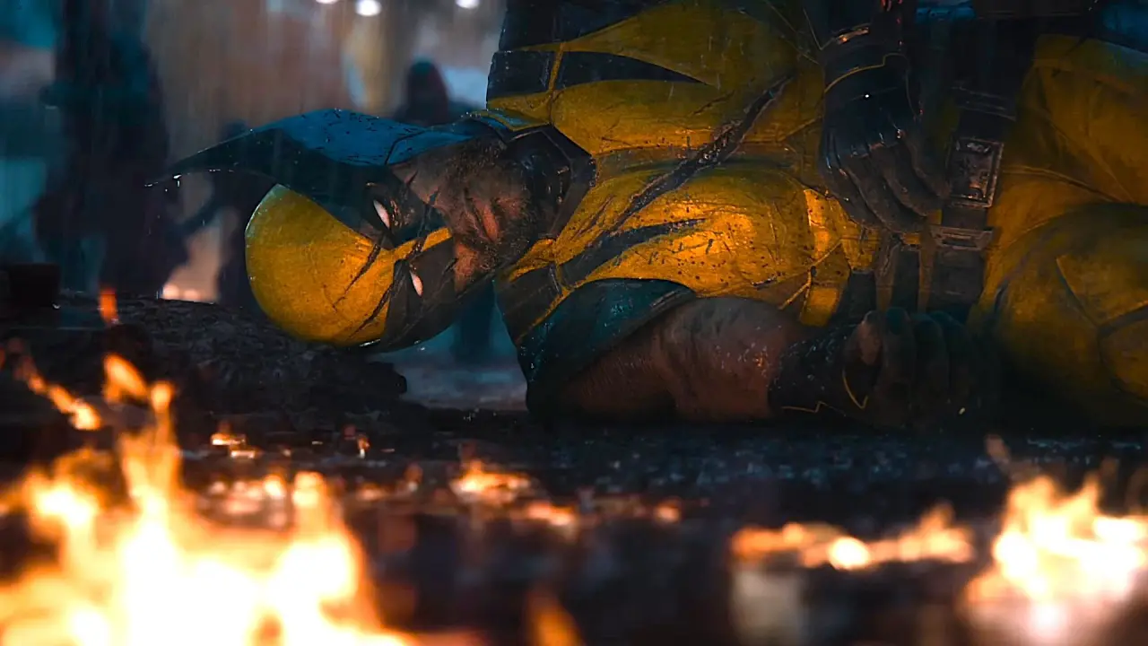 A character in a yellow and black costume lies on the ground, surrounded by flames, with a serious expression against a dark background.